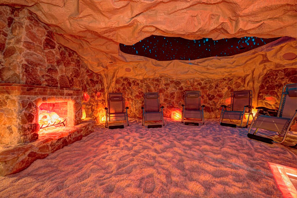An interior view of a salt room, featuring walls and a floor covered in pink and orange Himalayan salt. The floor is covered in fine, granular salt, giving it a bright, soft orange hue under the lighting. The walls are constructed with rough-hewn blocks of pink and orange salt, which are backlit to enhance their color and texture. In the center of the image, two black, reclining zero-gravity style chairs are positioned, facing forward. Each chair has a light-colored, possibly beige or white, towel draped over the backrest. The chairs have black mesh fabric seating and armrests. Between the chairs, set into the salt wall, is a recessed feature that resembles a window or fireplace. It has a dark, wooden frame and a lower section paneled with horizontal wooden planks, possibly acting as a mantle or storage. Inside the recessed area, the back wall is made of dark, textured rock or stone. A decorative arrangement of white, thin, branch-like elements, possibly artificial, is placed inside, and it is illuminated, creating a contrast with the dark stone background and suggesting a natural or rustic aesthetic. To the far left of the image, another section of the salt wall features a smaller, rectangular niche with an internal light source, emitting a warm, soft orange glow, adding to the room's ambiance. The overall lighting in the room is warm, predominantly orange and pink, emphasizing the therapeutic and relaxing atmosphere of the salt cave.