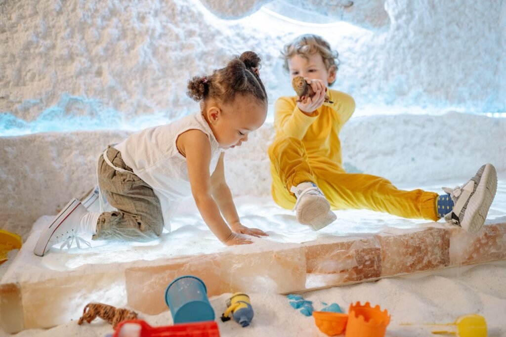 Two young children play joyfully in a salt room filled with toys and light.